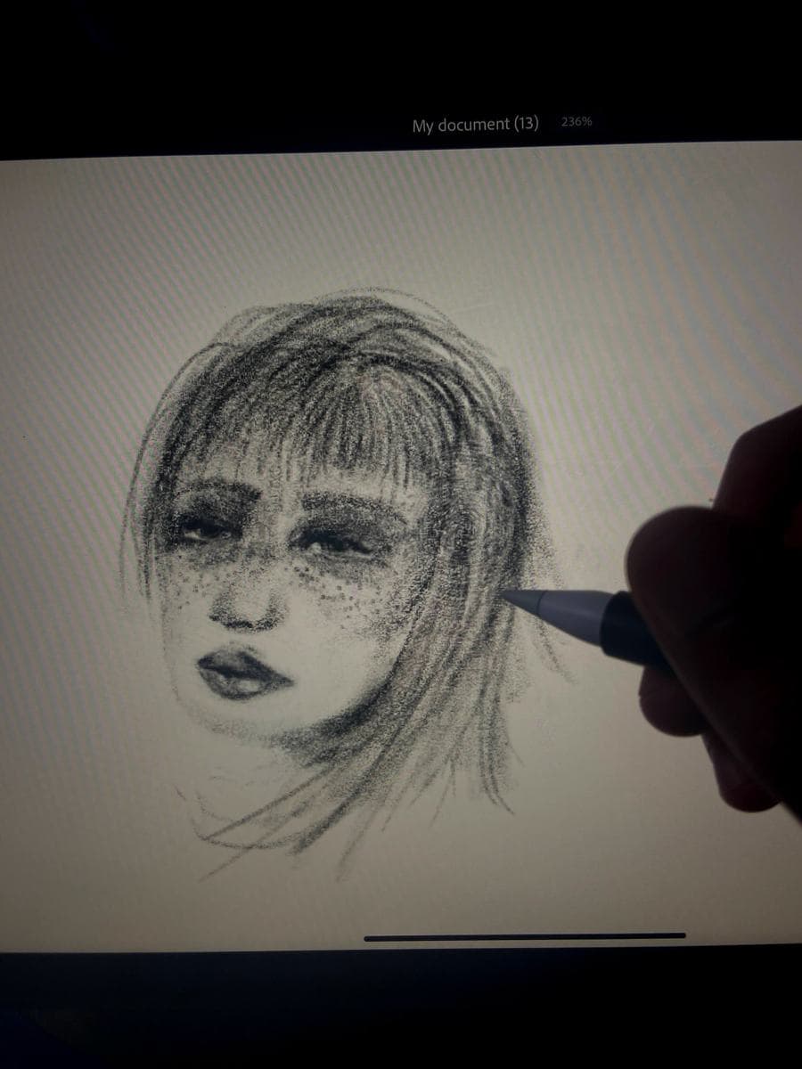 A tablet held in one hand, stylus resting on the screen, showing a soft charcoal portrait of a woman with a heavy fringe.