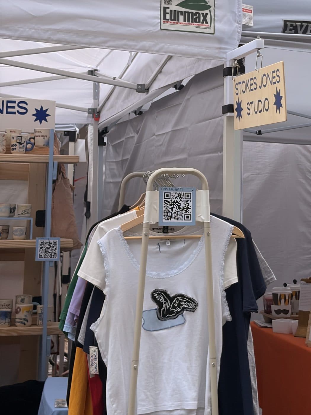 Two hand-lettered wooden shingles hung at right angles from a tent frame, with a QR card on a clothing rack.