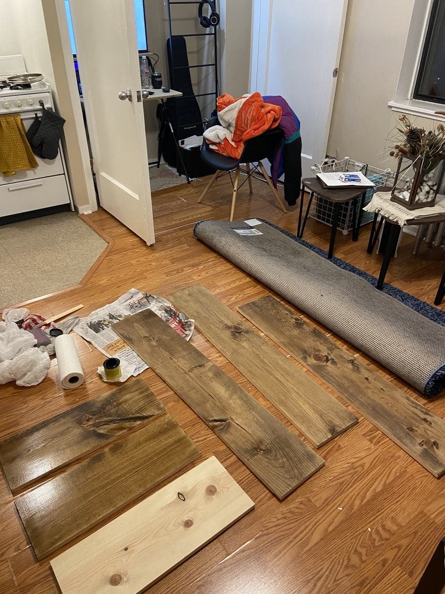 The boards laid out across a wood floor over spread newspaper, mid-job: five already stained, one still pale raw pine, with the tin, a roll of paper towel and used rags beside them.