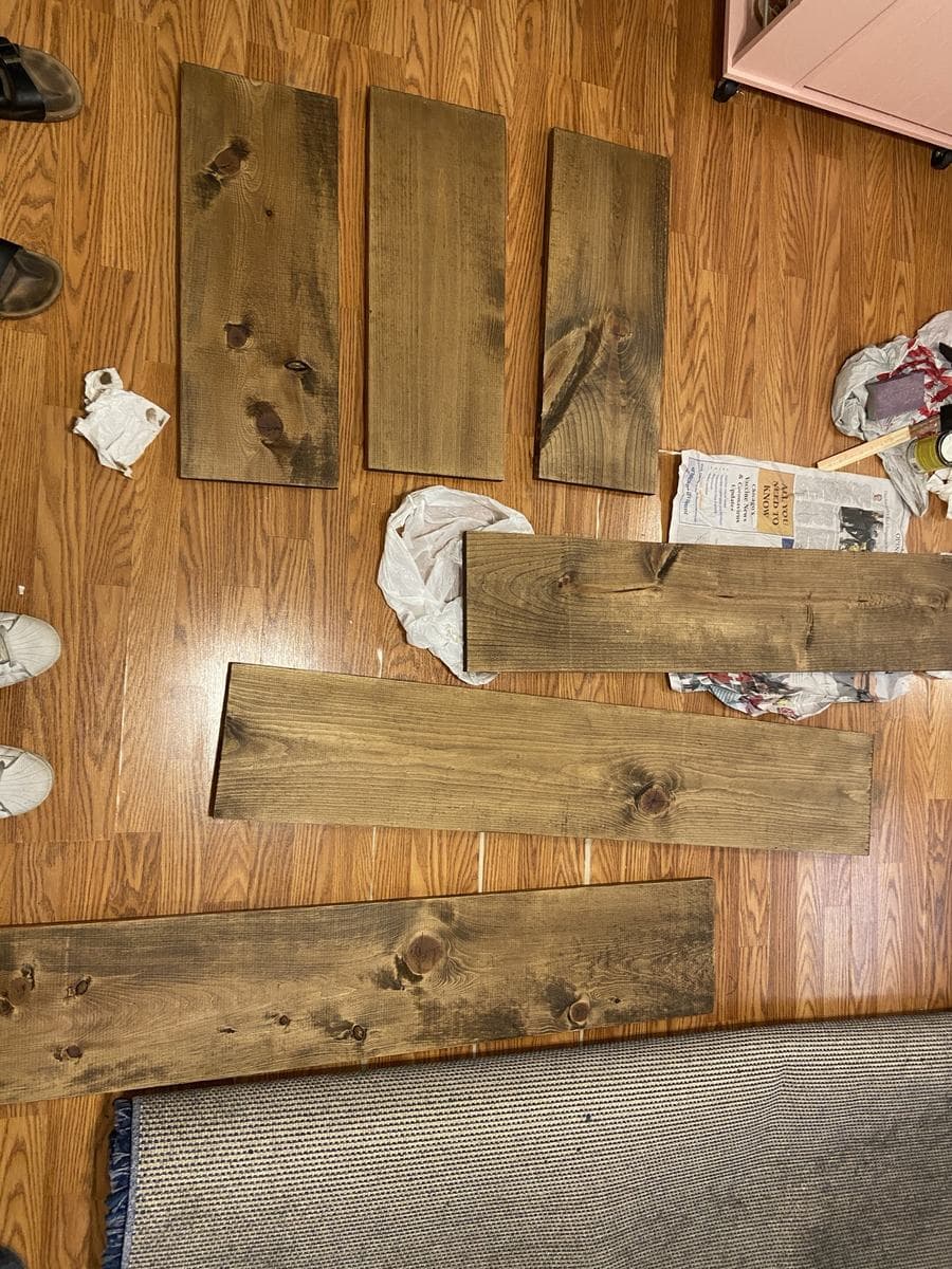 Six stained boards drying on the floor, seen from above: three short cross-braces in a row at the top and three long boards below them, with used rags, newspaper and the tin pushed to one side.