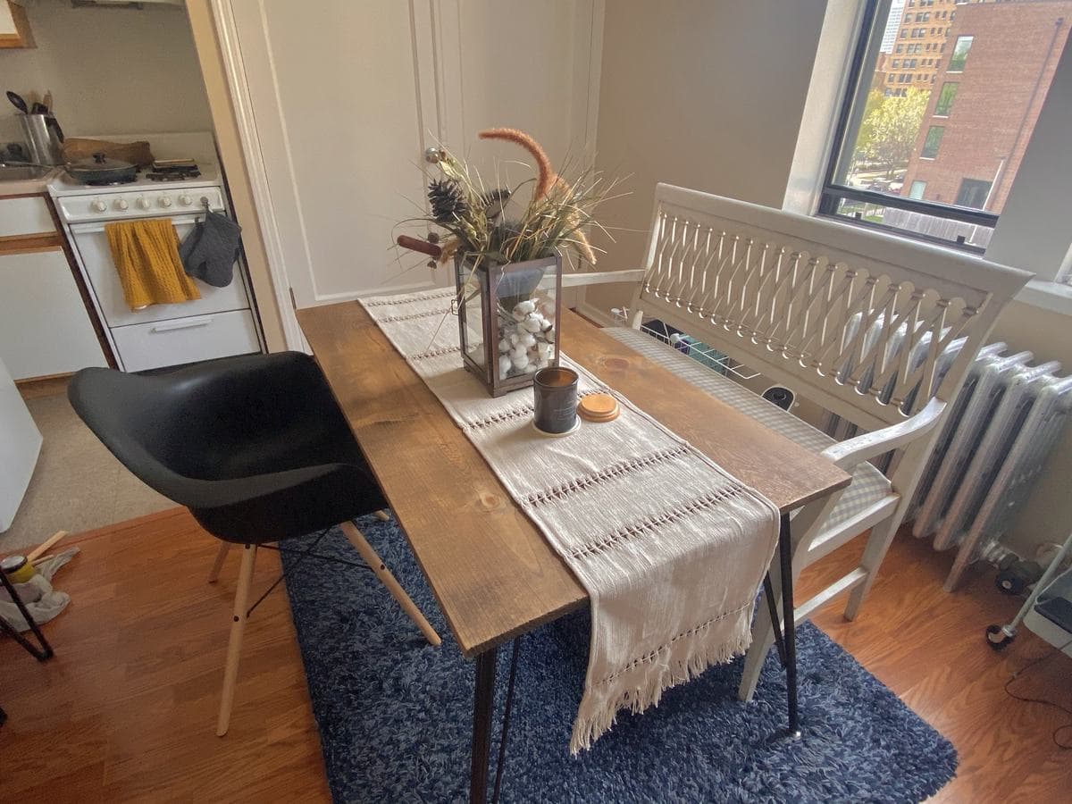 The finished table in the kitchen corner, dressed with a woven runner, a lantern of cotton bolls and a candle, a black moulded chair on one side and a white bench on the other.
