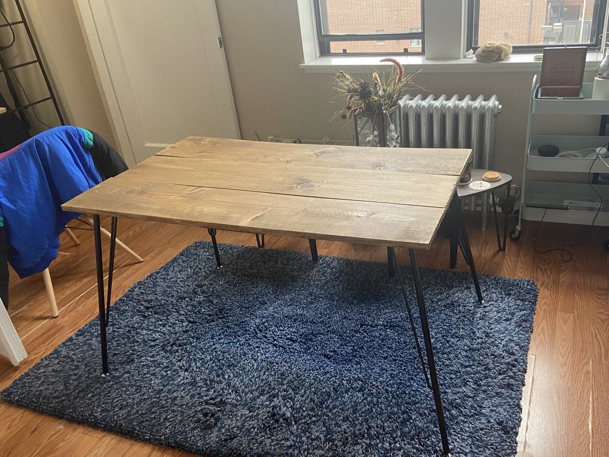 The same table from the other end, bare, standing on a blue shag rug on black hairpin legs, with a radiator and a window behind it.
