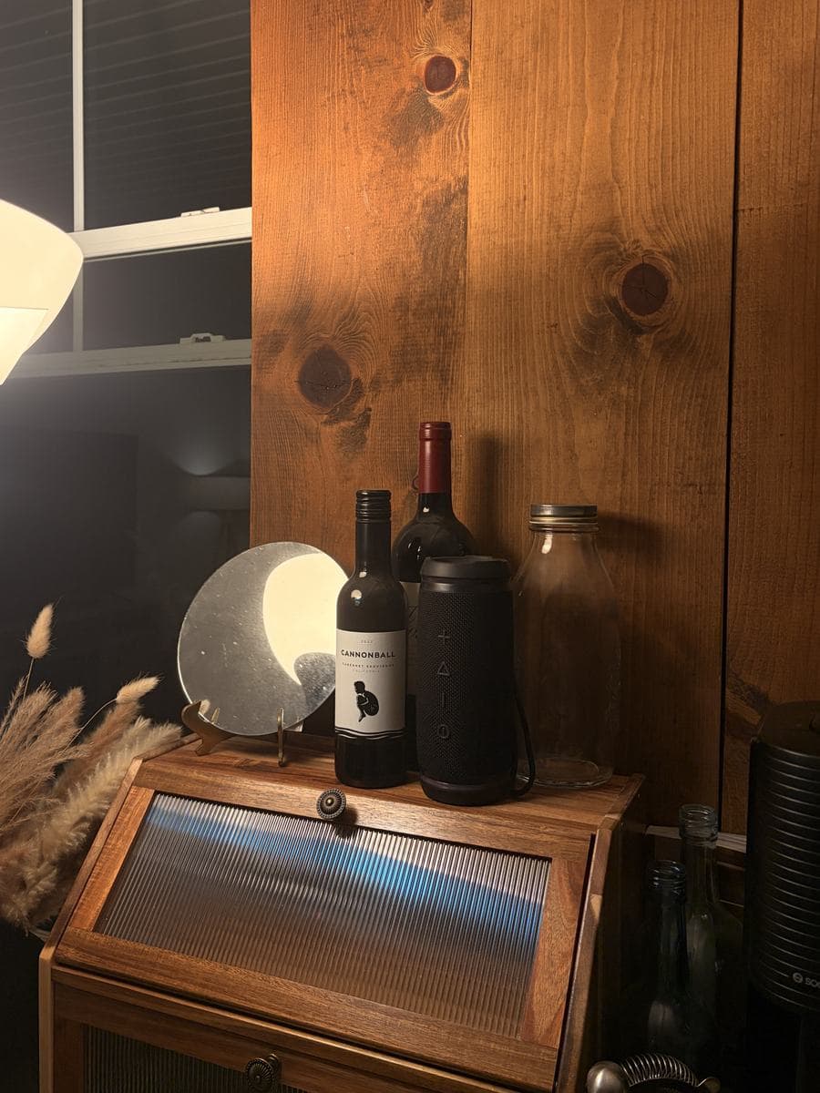The tabletop years later, mounted upright on a wall as a backdrop and lit warm by a lamp, standing behind a wooden cabinet topped with wine bottles, a speaker, a glass jar, a round stone disc on a stand and dried grasses.